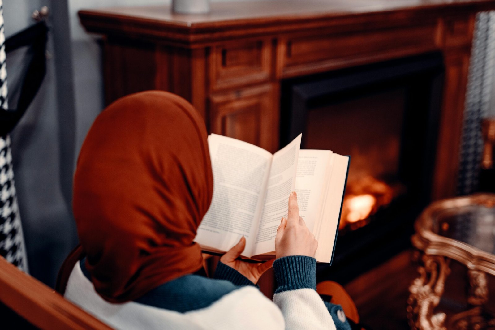 A woman in hijab reading a book by a warm fireplace, embracing a cozy and serene atmosphere.