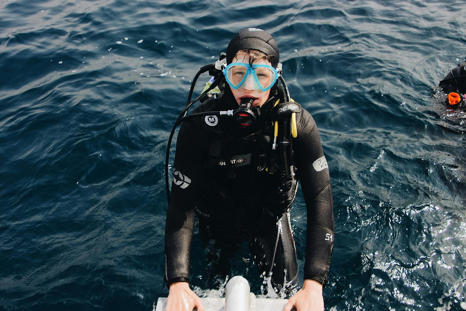 A scuba diver steps onto a boat ladder, emerging from the ocean, wearing full scuba gear.
