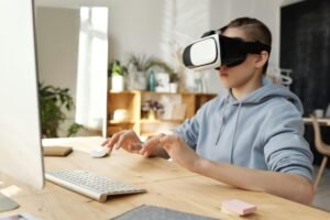 Teenager in gray hoodie using a VR headset indoors, sitting at a table with a keyboard and mouse.