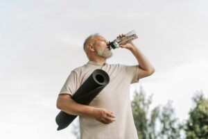 Elderly man drinking water while holding a yoga mat outdoors, promoting fitness and hydration.
