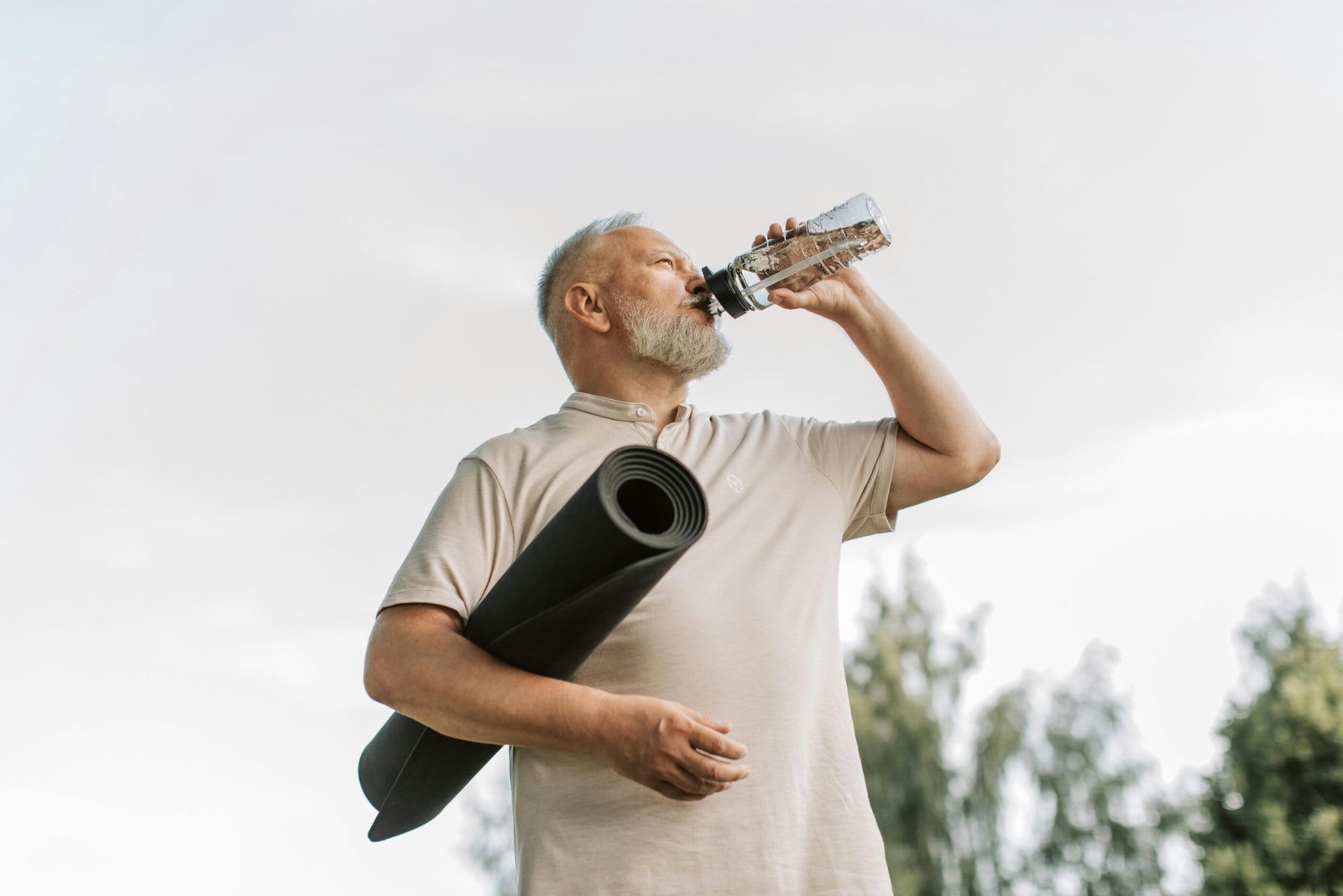 Elderly man drinking water while holding a yoga mat outdoors, promoting fitness and hydration.