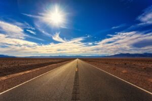 A long, straight road stretches across a dry desert landscape under a bright blue sky and shining sun.