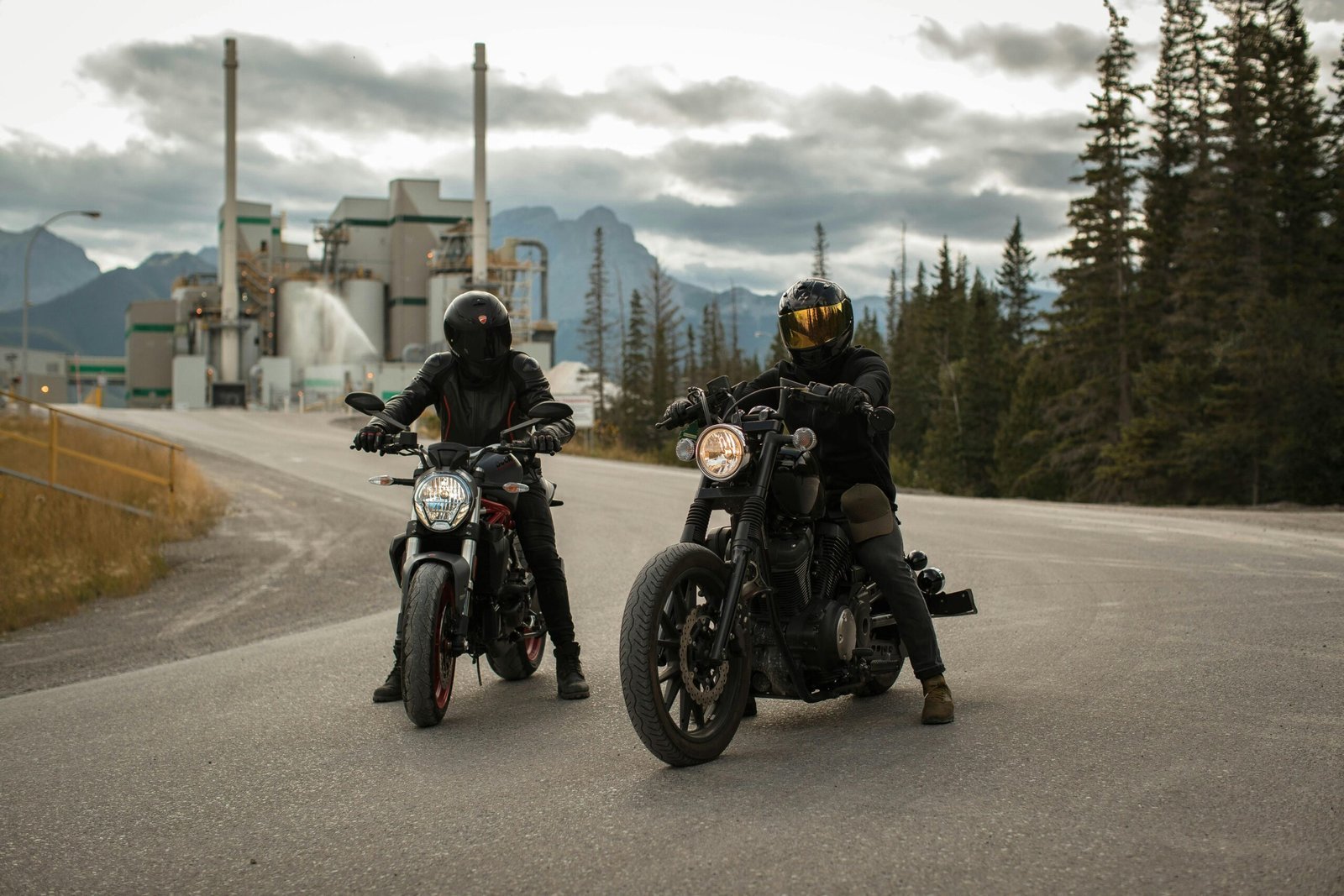Two motorcyclists pause on a road with a factory and forest backdrop in Canada.
