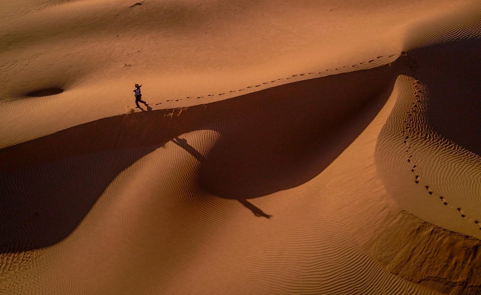 A lone person walks across sand dunes at sunset, casting long shadows in the desert.