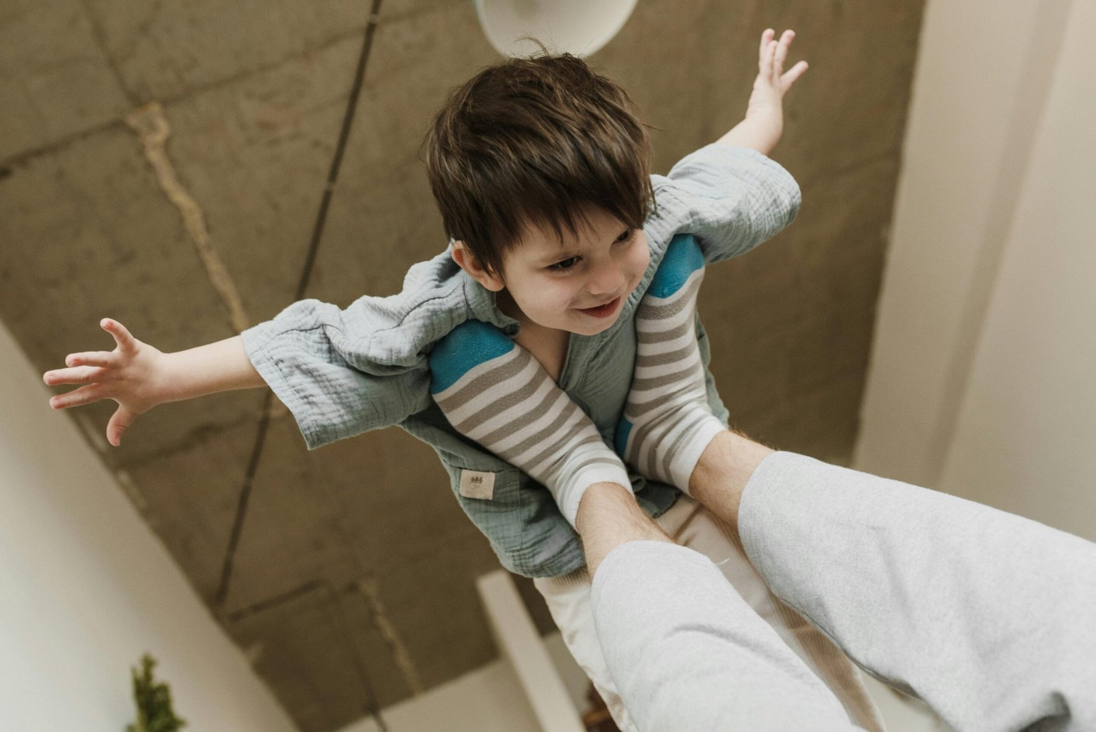 A smiling child enjoys a playful airplane game indoors, lifted by adult feet.