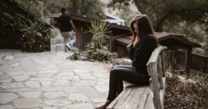 A woman sits pensively on a bench outside after a breakup, while a man walks away.