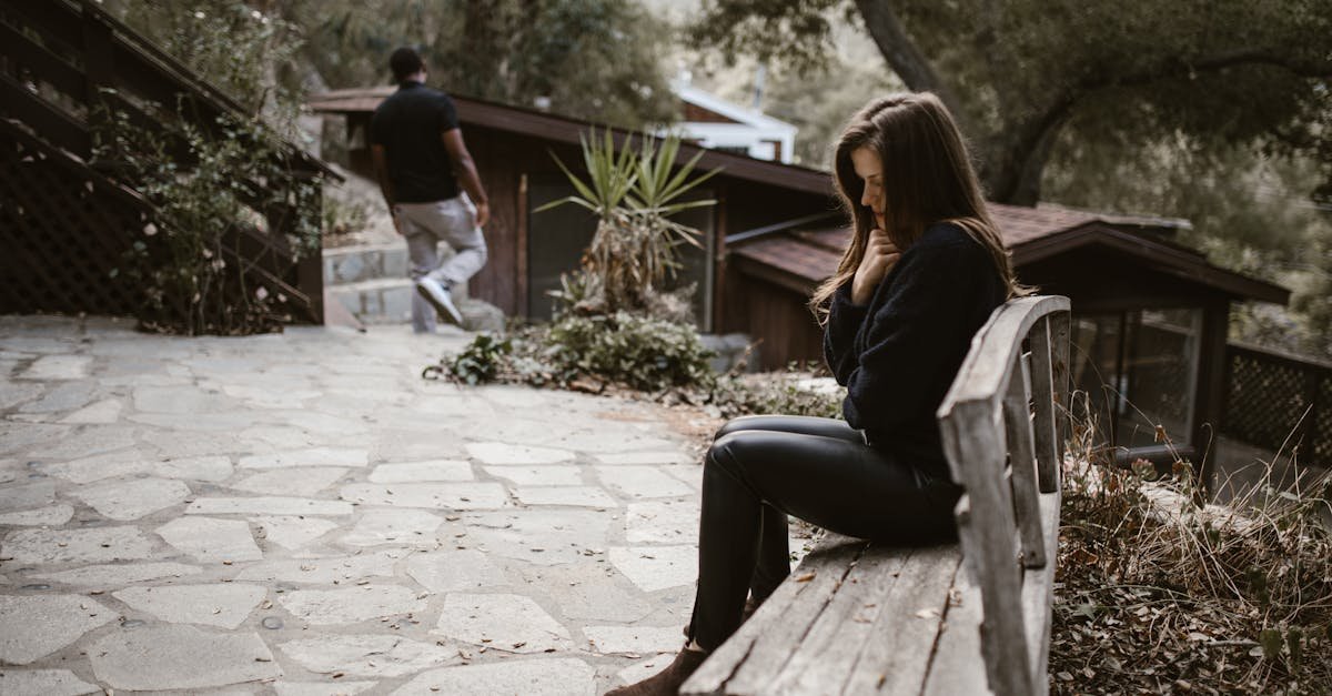 A woman sits pensively on a bench outside after a breakup, while a man walks away.