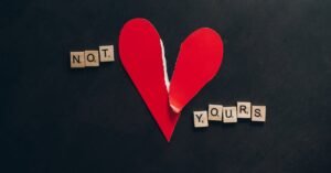 Artistic image of a torn red heart with scrabble tiles spelling 'Not Yours' on a black background.