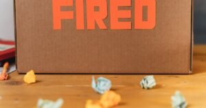 Close-up of a cardboard box labeled FIRED on a wooden table with crumpled papers.