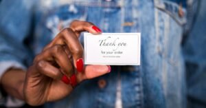 A woman in a denim jacket holds a 'Thank You' card with red nails.