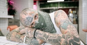 Tattooed man wearing bandana, lying in modern, tiled space with artistic decor.