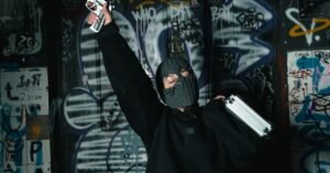 A masked figure in black sweater with gun and briefcase stands in a graffiti-covered alley.