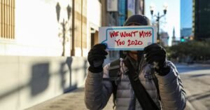 A person in winter clothing holds a sign reading 'We Won't Miss You 2020' on a city street.