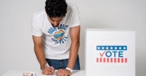 A man writes on a paper to cast his vote next to a ballot box in a studio setting.
