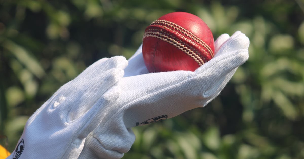 Detailed photo of a red cricket ball held by gloved hands outdoors.